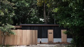 A modern building with a wooden exterior, a child playing in the yard, and dense foliage in the background. - Negura (Ogimi)