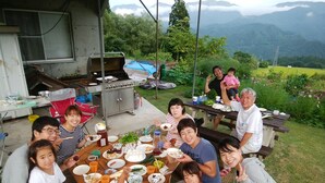 A group of people gathered around a wooden table outdoors with a grill and various food items. - Pension Silver Fern (Otari)
