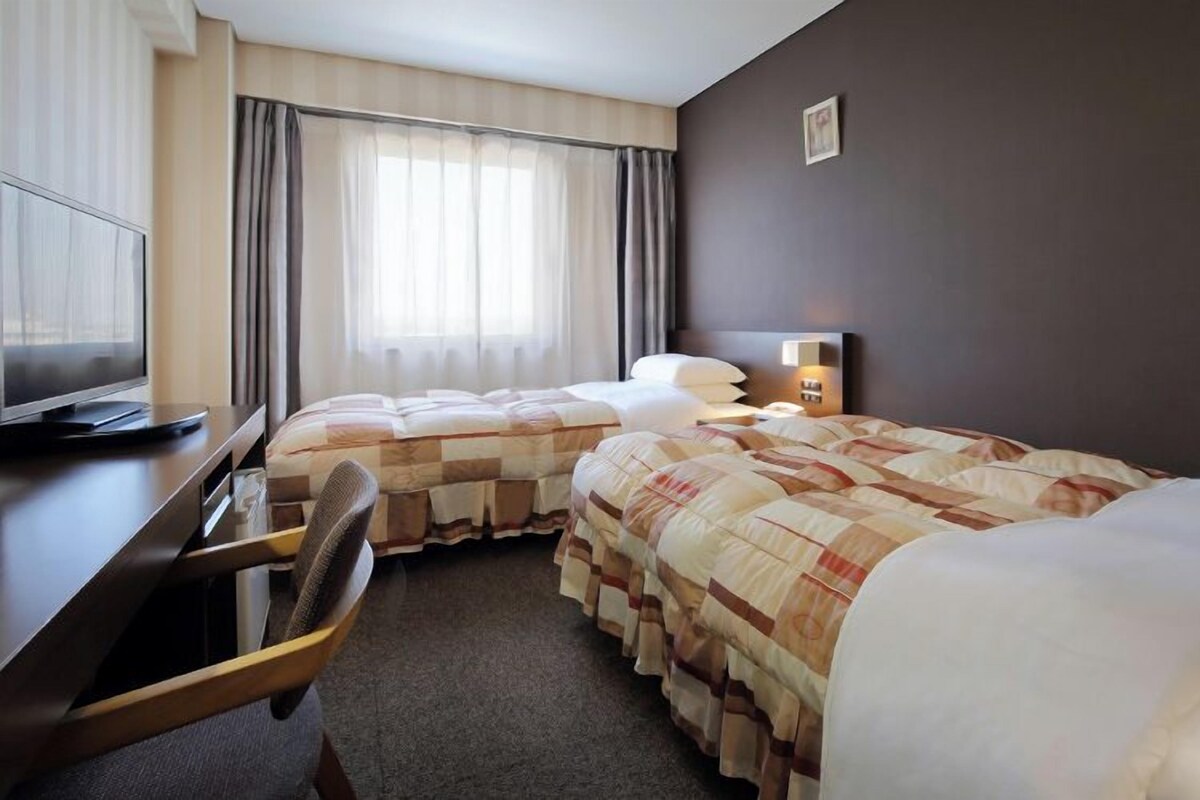 A hotel room with two beds, a desk, a chair, a TV, and a window with curtains.