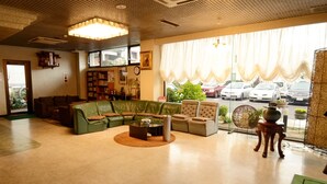 A spacious living room with a green sofa, a round coffee table, and a beige armchair. There is a large window with curtains, a bookshelf, and a chandelier. - Hotel Irihama (Sakaide)