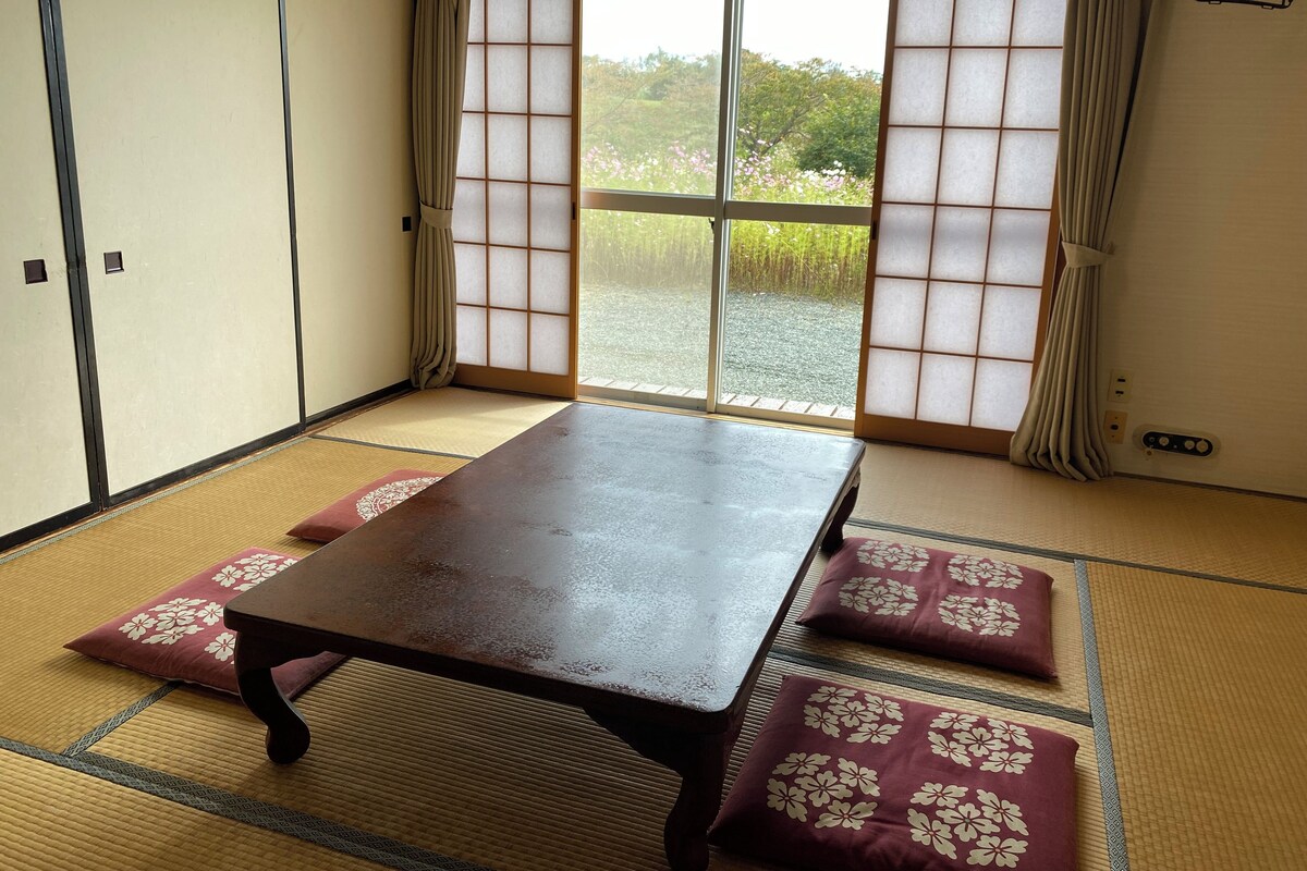 A tatami mat room with a wooden table, cushions, sliding shoji doors, and a view of greenery outside.