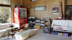 A room with a sofa, a table with a vase, a bookshelf, a Coca-Cola vending machine, and a window with a view of trees. - Edogawa Garden (Iwaki)