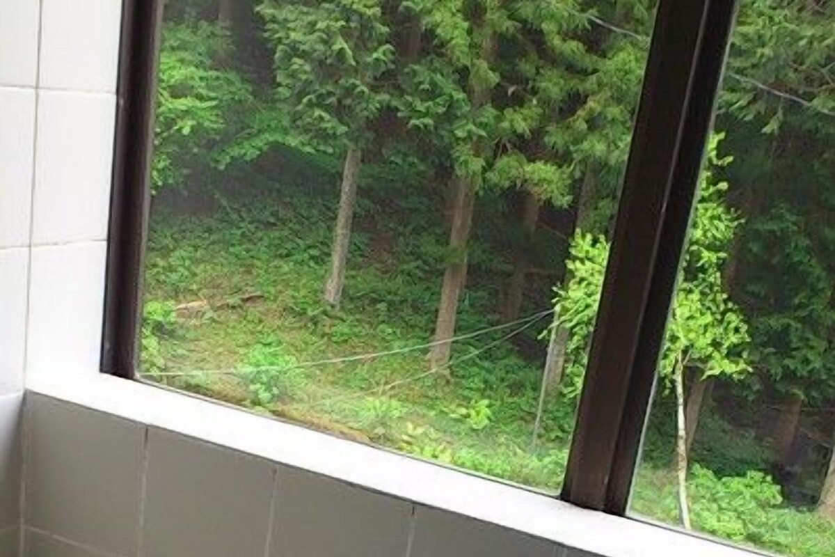 A bathroom with a bathtub, a window with a view of a forest, and a tiled floor.