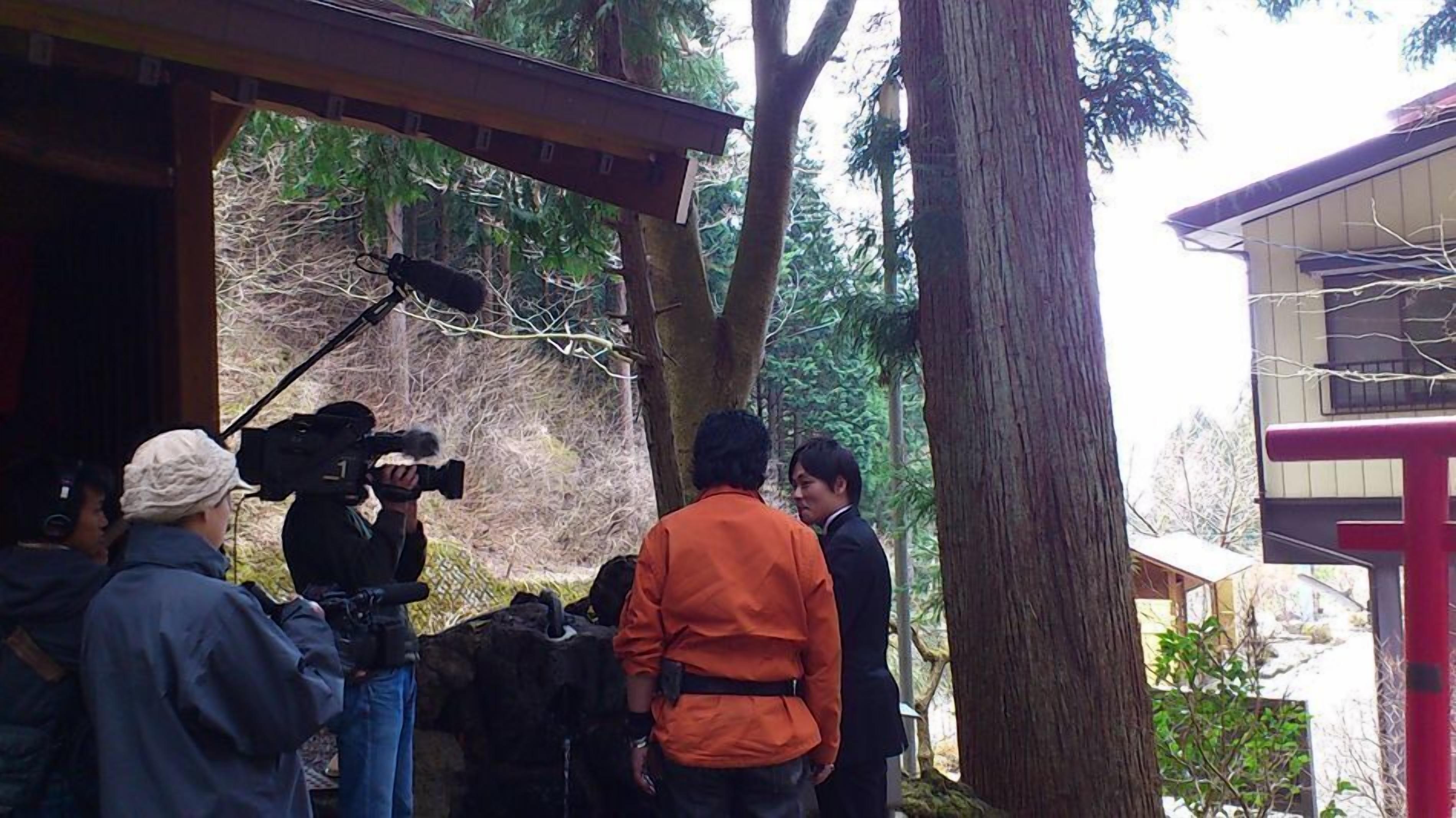 A group of people around a camera, possibly setting up for a photo or video shoot in a wooded area with a building in the background.