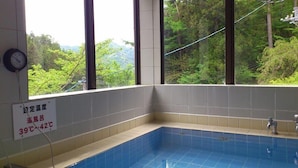Un coin de pièce avec une piscine aux carreaux bleus, un thermomètre indiquant une température de 39 à 42 °C, et une fenêtre donnant sur un paysage verdoyant.