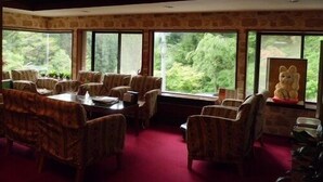 Un espace repas comprenant une grande table et des chaises, de grandes fenêtres donnant sur un paysage verdoyant et un mur en pierre.