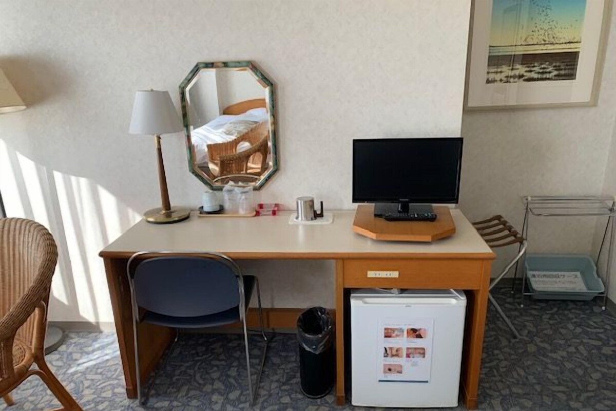 Ein Hotelzimmer mit einem Holztisch, einem Computer, einem Spiegel, einem Stuhl, einem kleinen Kühlschrank und einer Mülltonne.