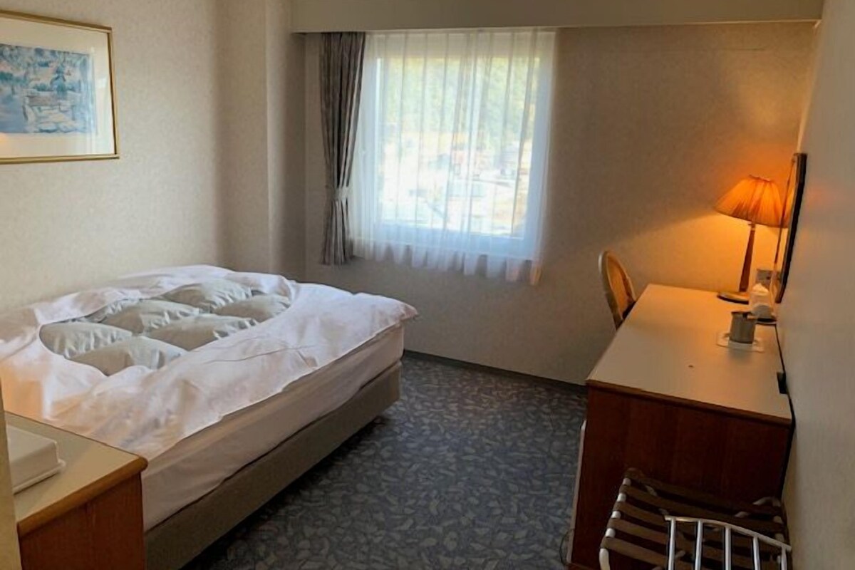Ein Hotelzimmer mit Bett, Schreibtisch, Stuhl, Lampe, einem Fenster mit Vorhängen und einem Bild an der Wand.