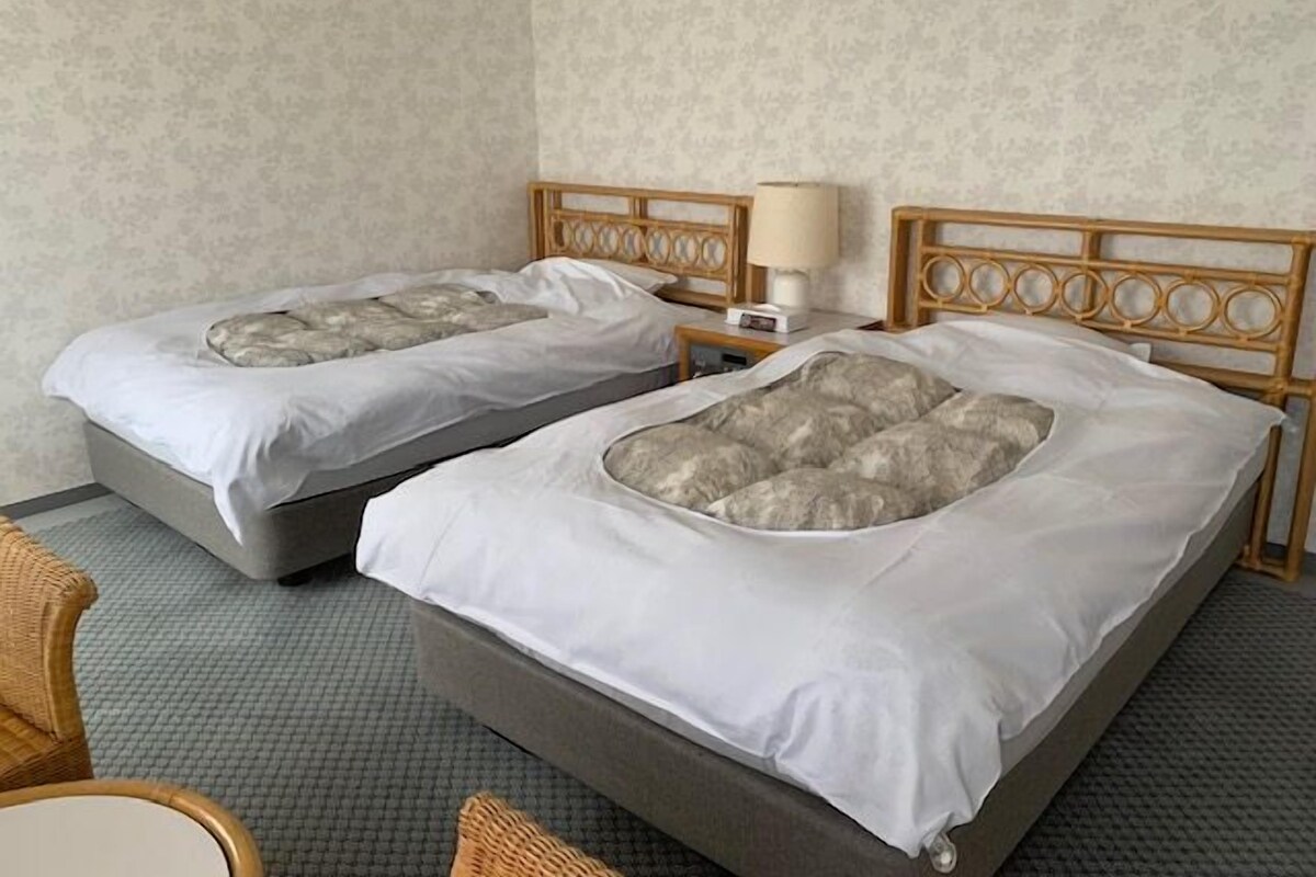 Dos camas individuales con ropa de cama gris y cabecero de madera, una mesita de noche con lámpara, y una silla de mimbre.