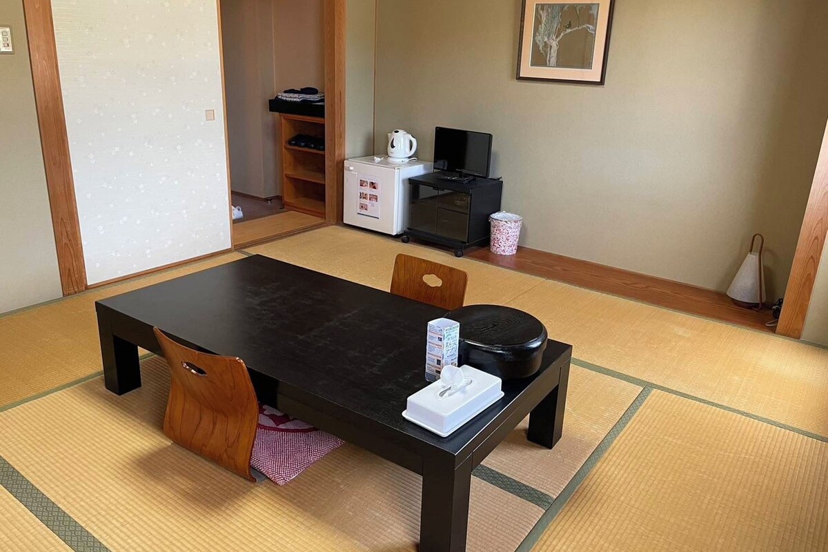 Une pièce de style japonais traditionnel, avec un sol recouvert de tatamis, une table basse et un meuble pour la télévision, ainsi qu’une bouilloire et un petit réfrigérateur.