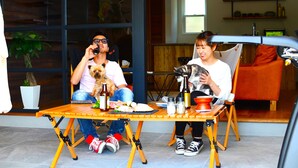 Deux personnes sont assises à une table en bois, accompagnées d’un chien, d’une bouteille et de divers aliments.