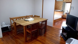 A room with a wooden dining table, four chairs, a television on a stand, and a microwave on a small table. - Amami no Yado Good Green (Setouchi)