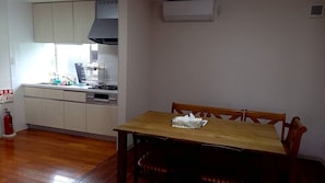 A dining area with a wooden table and chairs, a kitchen with white cabinets and a stove, and a fire extinguisher on the floor. - Amami no Yado Good Green (Setouchi)
