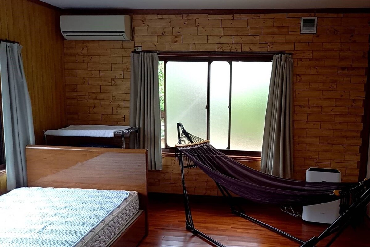 Una habitación con cama, una hamaca, una mesita y una ventana con cortinas.