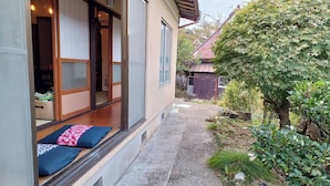A traditional Japanese-style building with a sliding door, a small outdoor seating area with cushions, and a garden with various plants and a tree. - Moritasanchi (Ine)