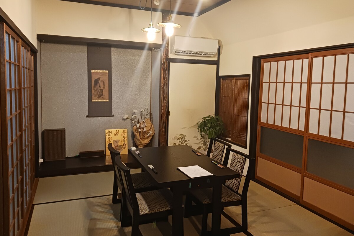 A traditional Japanese room with tatami flooring, a low table with chairs, a shoji screen, and a mounted air conditioner.