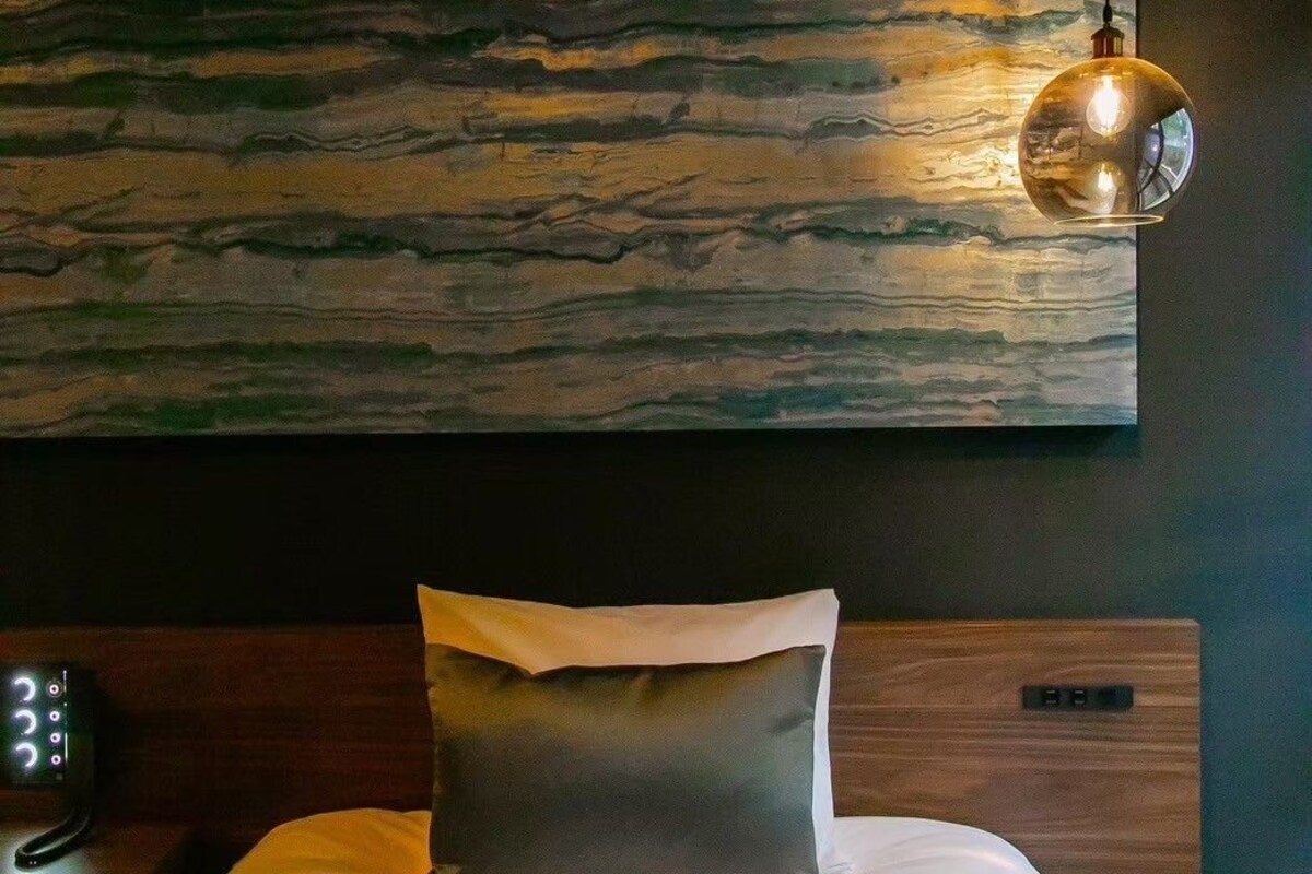 Una cama bien tendida con un almohadón, una lámpara de pared y un panel de control a la izquierda.