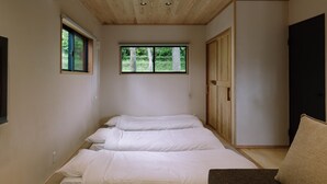 Ein Schlafzimmer mit Holzbalkendecke, einem Fenster mit Blick auf Bäume, einem Bett mit weißer Bettwäsche und einem dunkelgrauen Sofa.