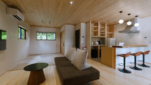 Ein modernes Wohnzimmer mit Holzbalkendecke, einer Couch, einem Essbereich mit Barhocker und einer Küche mit integriertem Kühlschrank.