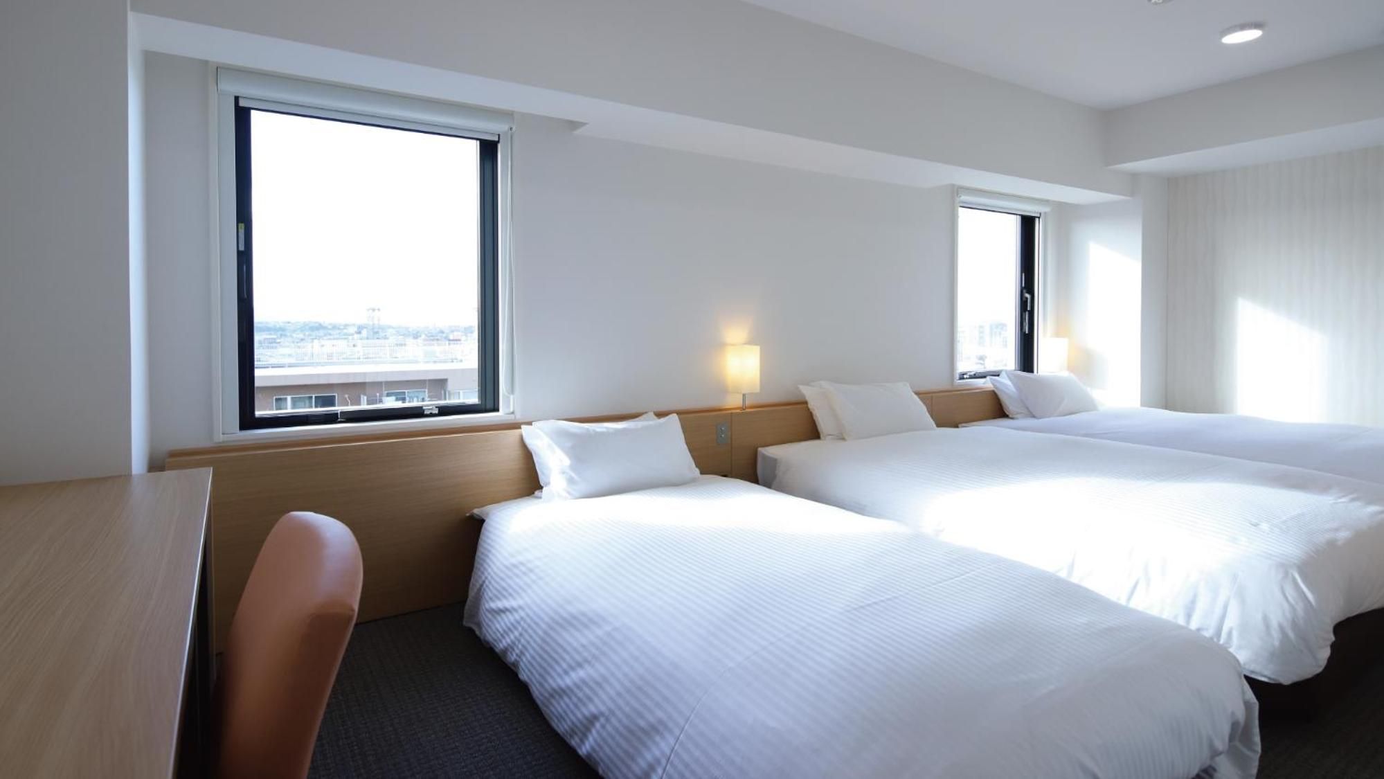 A hotel room with two beds, a desk, and a chair. There are windows providing a view of the city.