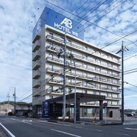 Un hotel de varios pisos con un llamativo letrero 'AB HOTEL', un área de estacionamiento y un paso de peatones.