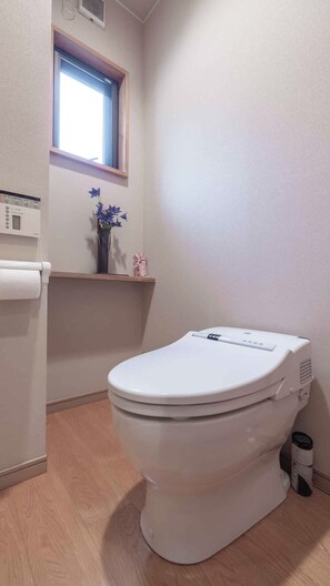 A bathroom with a toilet, a window, a small shelf with decorative items, and a wall-mounted heater. - Temple Hotel Matsui-bo (Minobu)