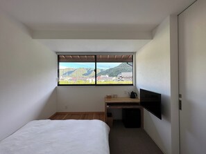 A small, modern room with a bed, a desk, a TV, and a window with a view of hills and buildings. - Bizen Hotel Sue (Bizen)