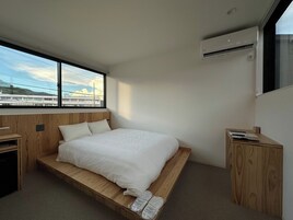 Un dormitorio moderno con una cama de madera, una mesita de noche pequeña y una ventana con vistas a edificios y líneas eléctricas.