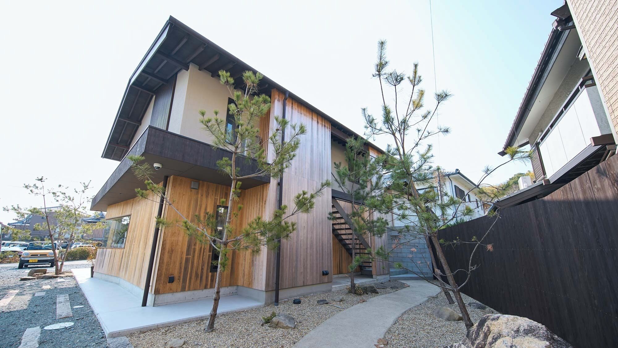 Una casa moderna de dos pisos con fachada de madera, una escalera visible y un pequeño jardín con grava y árboles jóvenes.