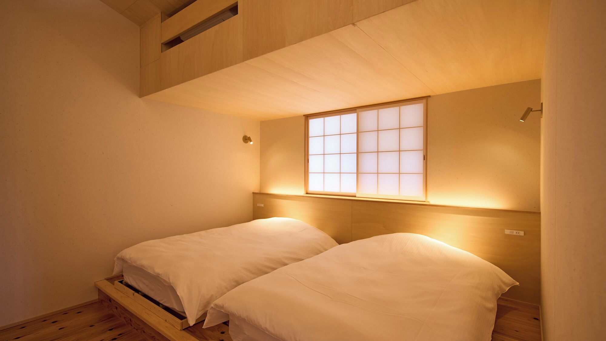 A room with two beds, a wooden headboard, a window with a grid pattern, and a small shelf above the beds.