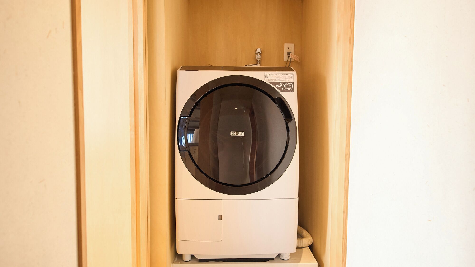 Une machine à laver compacte, de couleur blanche, à chargement frontal, intégrée dans un meuble en bois, dotée d’une porte circulaire, d’une prise de courant et d’un raccord d’eau situés sur le dessus.