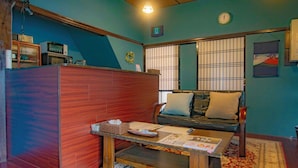 Ein Zimmer mit Holzwänden, einer Couch mit Kissen, einem Couchtisch mit Zeitschriften, einer Uhr an der Wand und einem Fenster mit Jalousien.