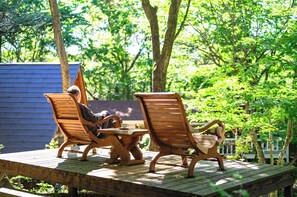 A wooden deck with two wooden chairs and a small table, surrounded by lush greenery. - Log Cottage Chausu no Mori (Nasu)