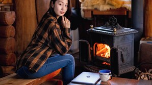 A person sitting by a wood-burning stove in a rustic room with wooden walls and a table with a laptop and mug. - Log Cottage Chausu no Mori (Nasu)