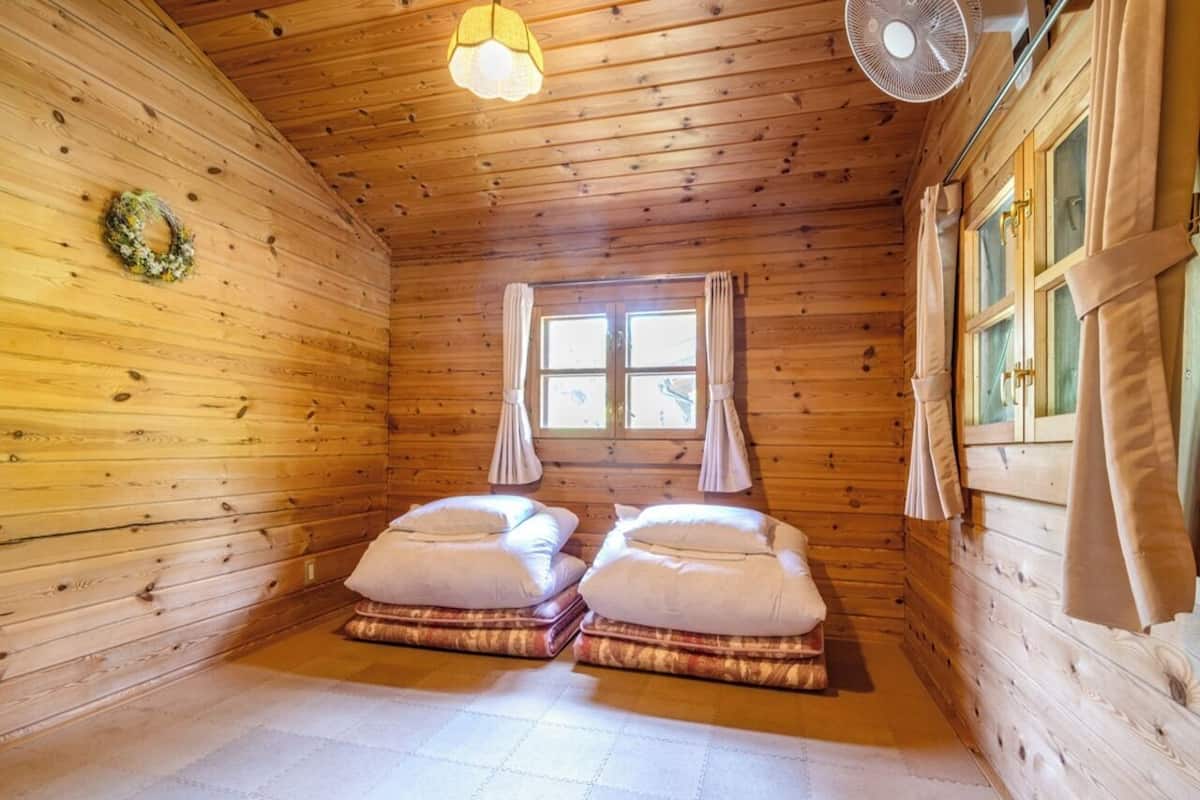 Una habitación de cabaña acogedora, con paredes de madera, dos camas con ropa de cama blanca, una ventana con cortinas y un ventilador de techo.