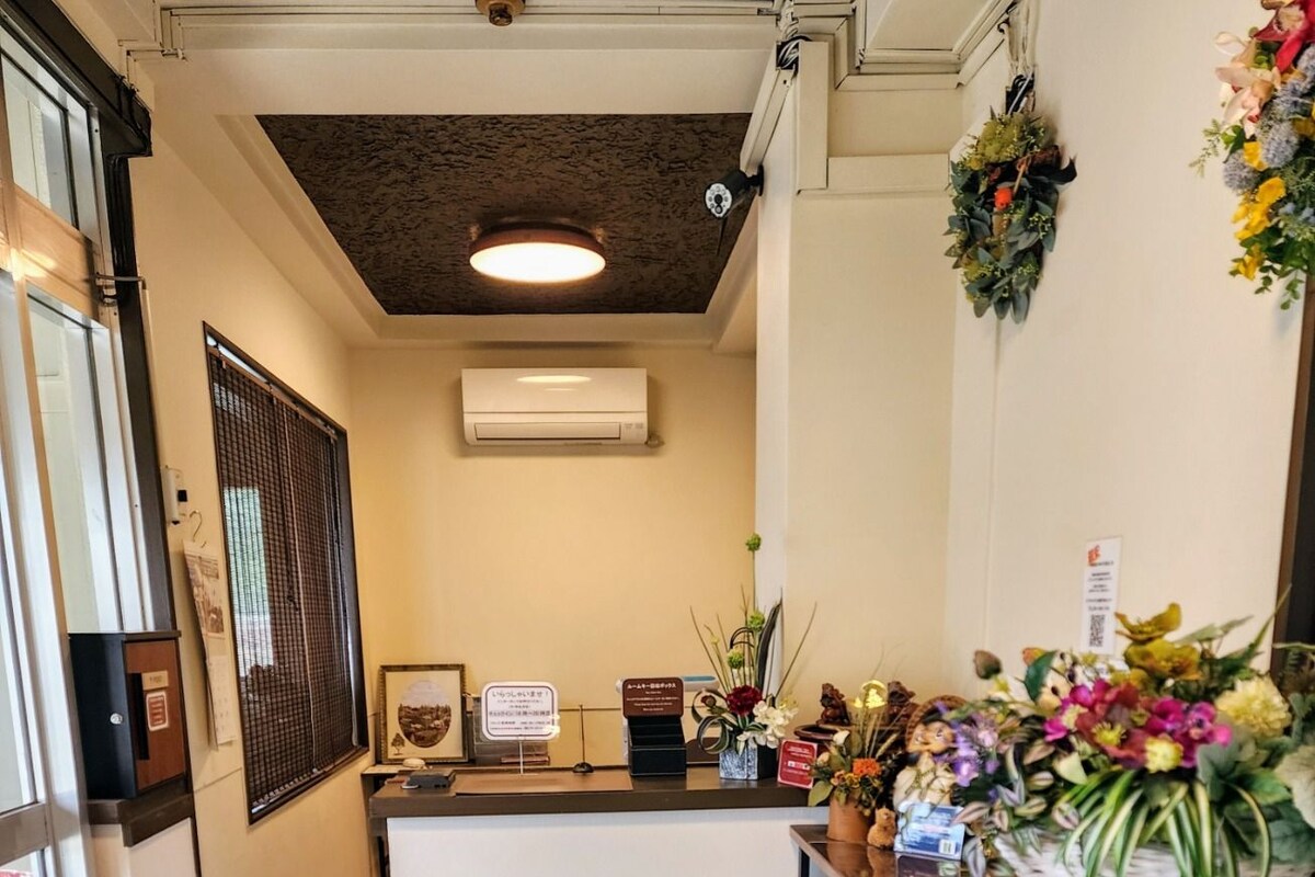 Ein Empfangsbereich mit einer hölzernen Bank, einem braunen Teppich, einem Empfangstresen mit einer Blumenkorb sowie einer wandmontierten Klimaanlage.