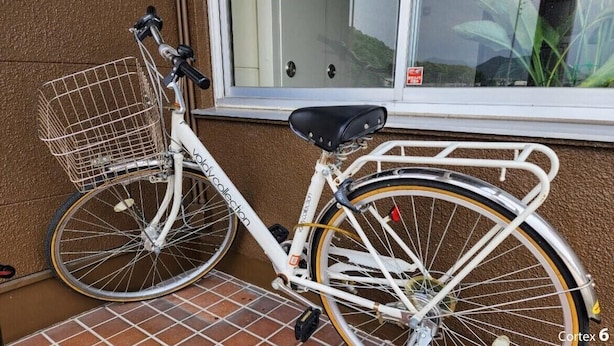 A white bicycle with a basket and a seat, parked against a wall with a window and a door in the background. - Business Hotel Nishiwaki (Nishiwaki)