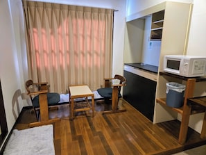 Une petite pièce, de taille modeste, avec un plancher en bois, deux chaises en bois, une petite table, un four à micro-ondes posé sur une étagère et une poubelle.