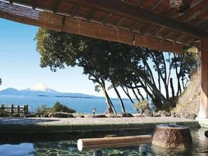 A tranquil outdoor pool area with a wooden deck, a stone wall, and a view of a mountain and the sea. - Benten Kousen (Minamiboso)