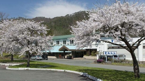 A hotel with a green roof, surrounded by cherry blossom trees, and a parking lot with cars. - Kanaya Onsen Hotel Ginga Park Hanamaki (Hanamaki)