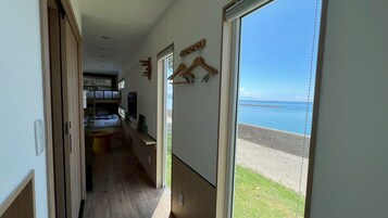 A narrow hallway leading to a bedroom with a bunk bed, a small round table, and a TV. There are wooden hangers on the wall and a view of the sea through a large window.
