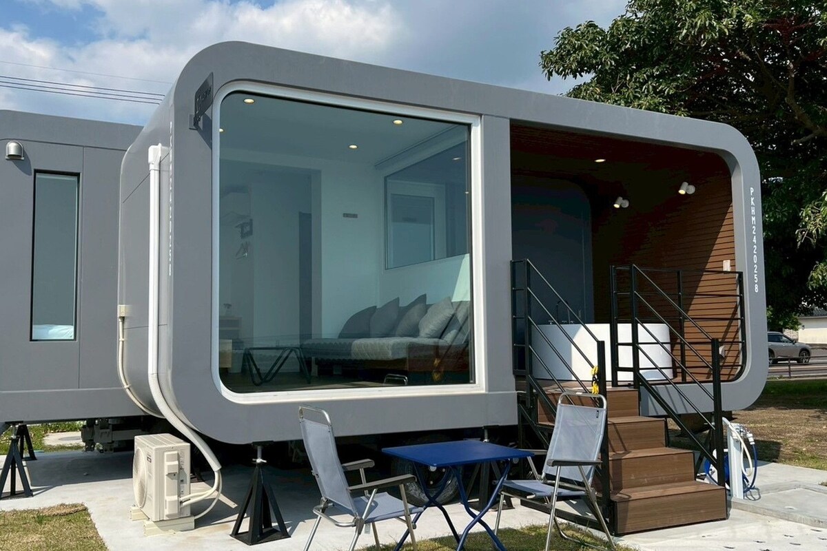 Una casa móvil moderna y compacta con un amplio ventanal, un pequeño porche y una escalera que lleva a un nivel superior.