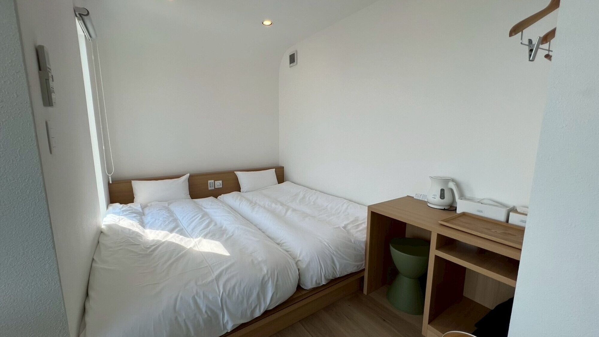 Una habitación pequeña y limpia con una cama individual, una mesita de noche de madera, un cubo de basura verde y una tetera blanca.