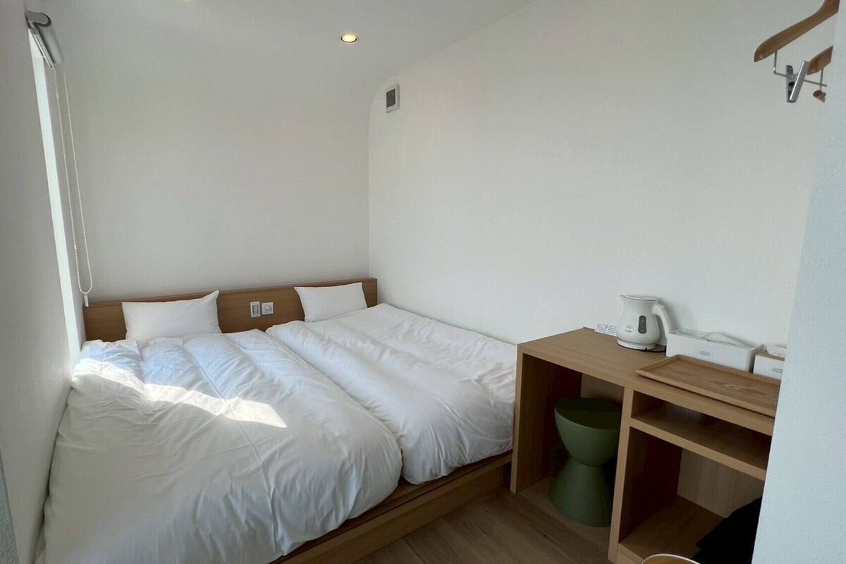 Una habitación pequeña y limpia con una cama individual, una mesita de noche de madera, un cubo de basura verde y una tetera blanca.
