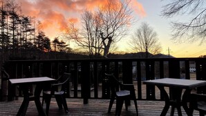 A wooden deck with tables and chairs, overlooking a forested area with a sunset in the background. - Kids Pension Maple (Hokuto)