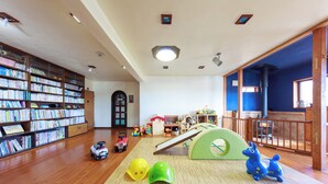 A spacious room with wooden flooring, a large bookshelf filled with books, a play area with a slide and a green tunnel, and a wooden railing overlooking a balcony. - Kids Pension Maple (Hokuto)