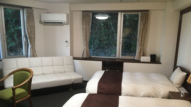Habitació d'hotel amb dos llits, un sofà, una cadira, un aparell d'aire condicionat i una finestra amb cortines.