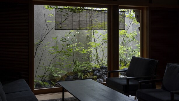 Een kamer met een schuifdeur die uitkomt op een tuin met rotsen en planten. In de kamer staat een donkergrijze bank en een klein tafeltje.