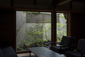 A room with a sliding glass door leading to a garden with rocks and plants. There is a dark-colored sofa and a small table in the room. - Toutou Achi no Sho Kura no Yado (Kurashiki)