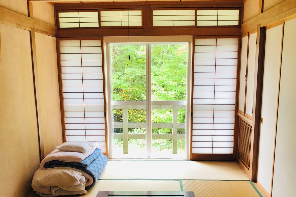 A traditional Japanese room with tatami flooring, a low table with a tea set, and a view of greenery through shoji screens.